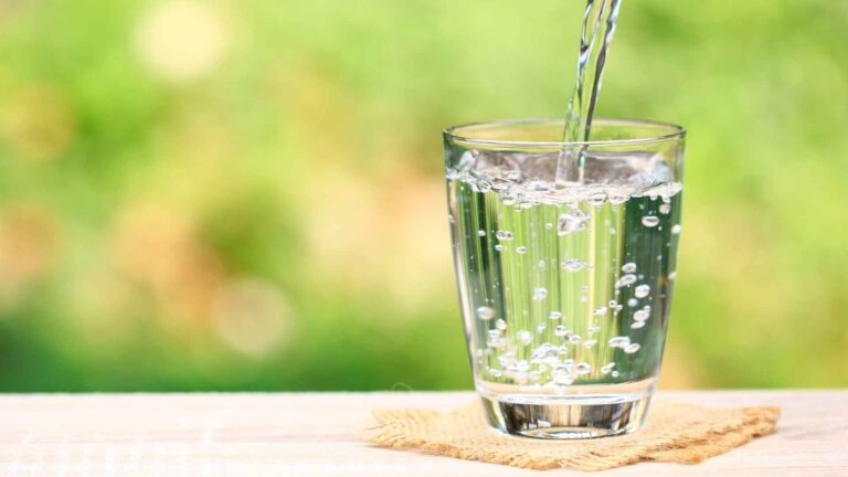 Clear water, reminiscent of solutions for water problems in Michigan, being poured into a glass on a wooden surface against a blurred green background.