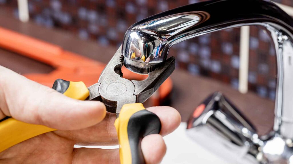 A plumber's hands using yellow and black pliers to adjust a shiny metal faucet over a kitchen sink.
