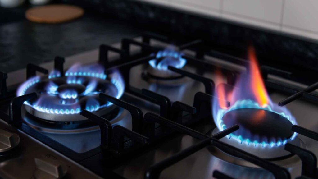 Gas stove burners lit with blue flames, one burner with a higher orange flame, set against a kitchen countertop featuring detailed kitchen plumbing.