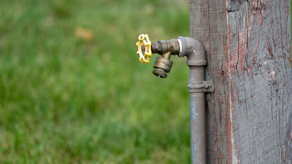 An outdoor plumbing fixture with a yellow handle mounted on a weathered wooden post, with a soft green background.