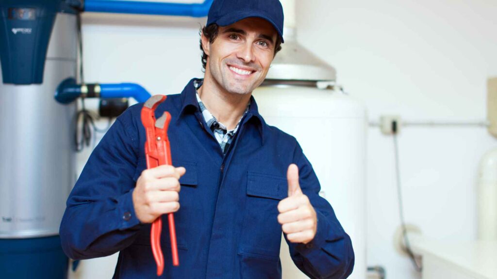 A smiling plumber in a blue uniform holding pliers and giving a thumbs up next to a sump pump in a boiler room.