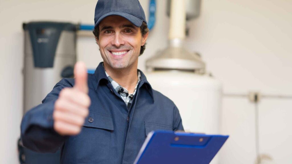 A smiling male technician in a cap and work uniform holding a clipboard and giving a thumbs up near a sump pump in a boiler room.