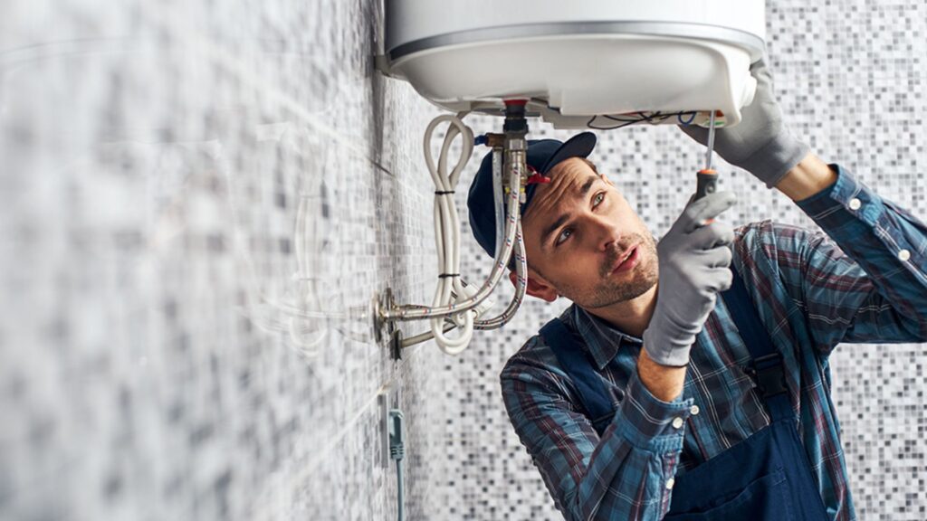 A plumber wearing a plaid shirt and overalls repairs a tankless water heater while looking thoughtfully at the connections.