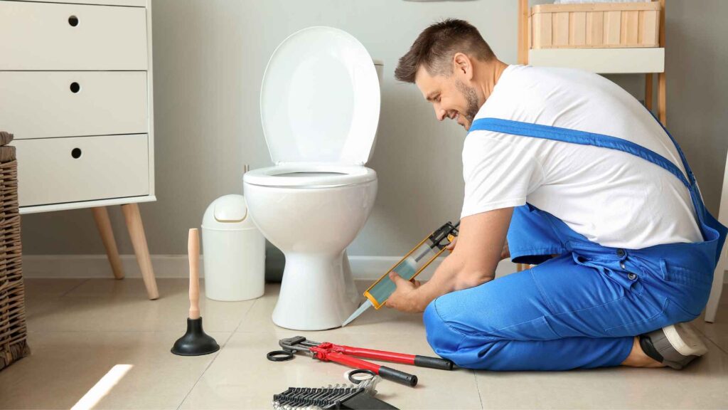 A smiling plumber in blue overalls fixing a toilet with bathroom plumbing tools around him on a bathroom floor.