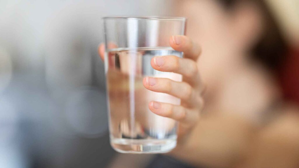 A person holding a clear glass of water to demonstrate water quality, extending it towards the camera with a blurred background.