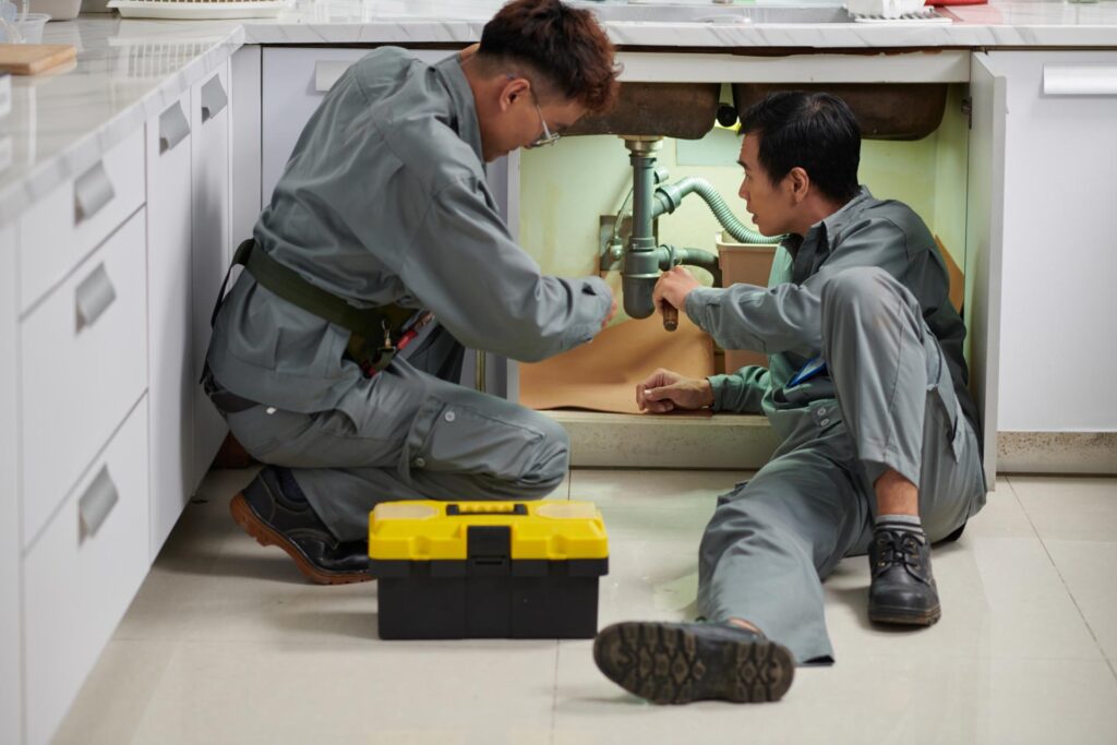 Two technicians inspecting and repairing home plumbing under a sink.