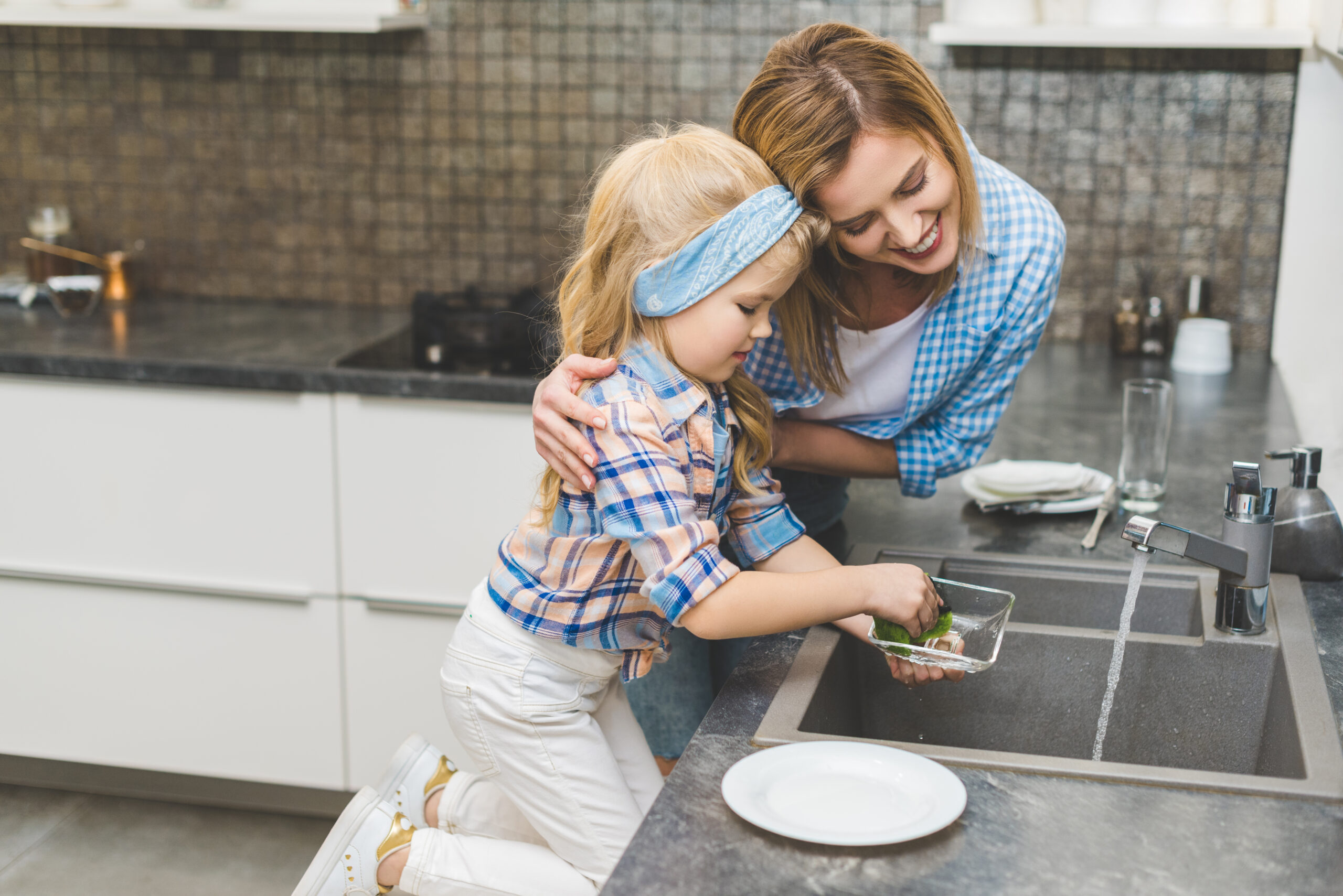 A woman and a young child, smiling and performing aspen maintenance on dishes together in a kitchen.