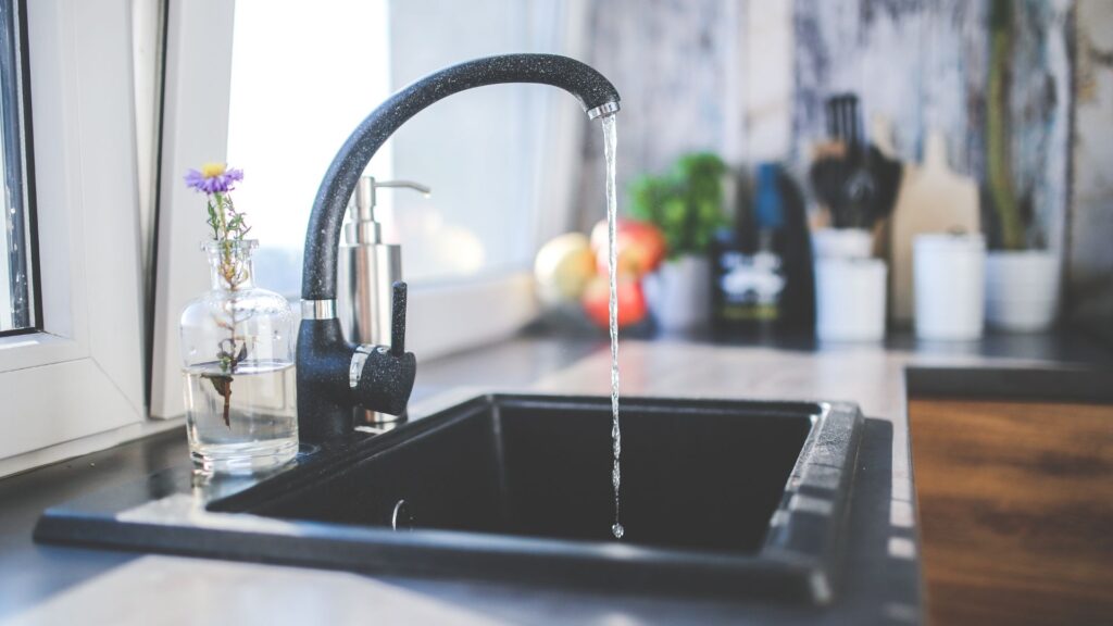Running water from a modern kitchen faucet into a black sink with kitchen items in the background.