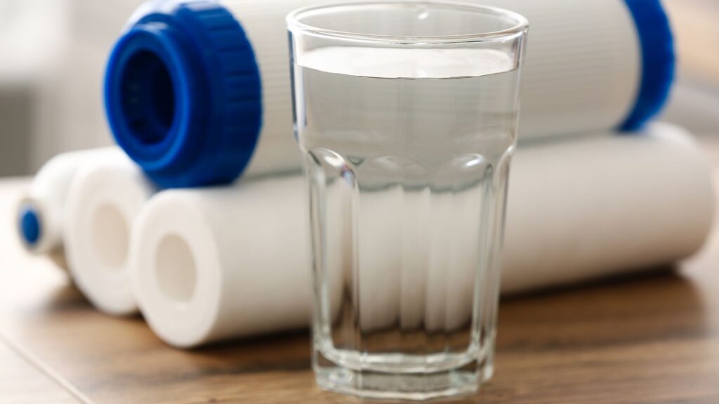 A glass of water in focus with a background of assorted water filters.
