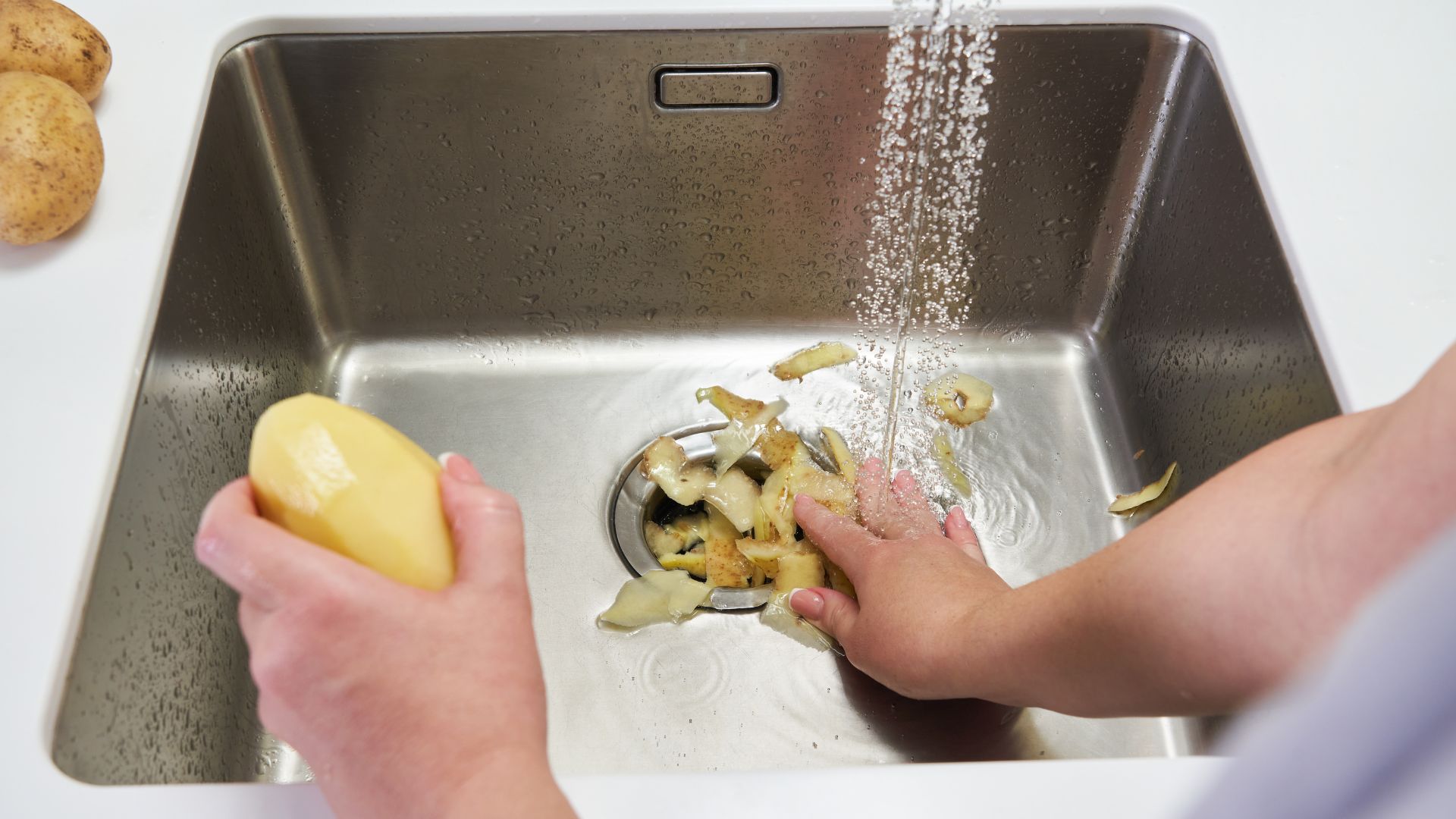 Person peeling potatoes over a kitchen sink and putting the peels in the garbage disposal
