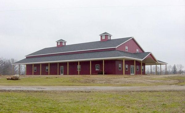A large red barn with a silver roof and white trim, featuring a covered porch area and twin cupolas, located in a flat, open field.