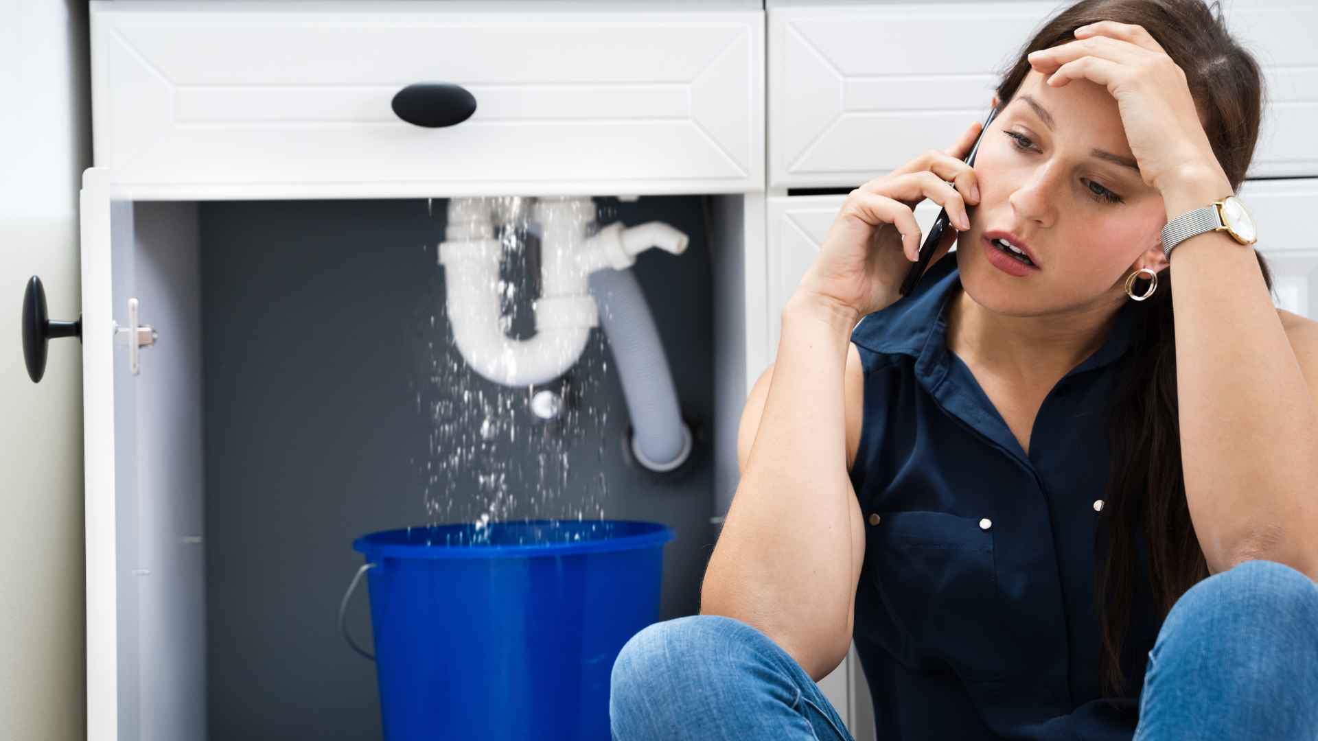 A woman on the phone, looking stressed, as hidden leaks drip from a sink pipe into a bucket below.