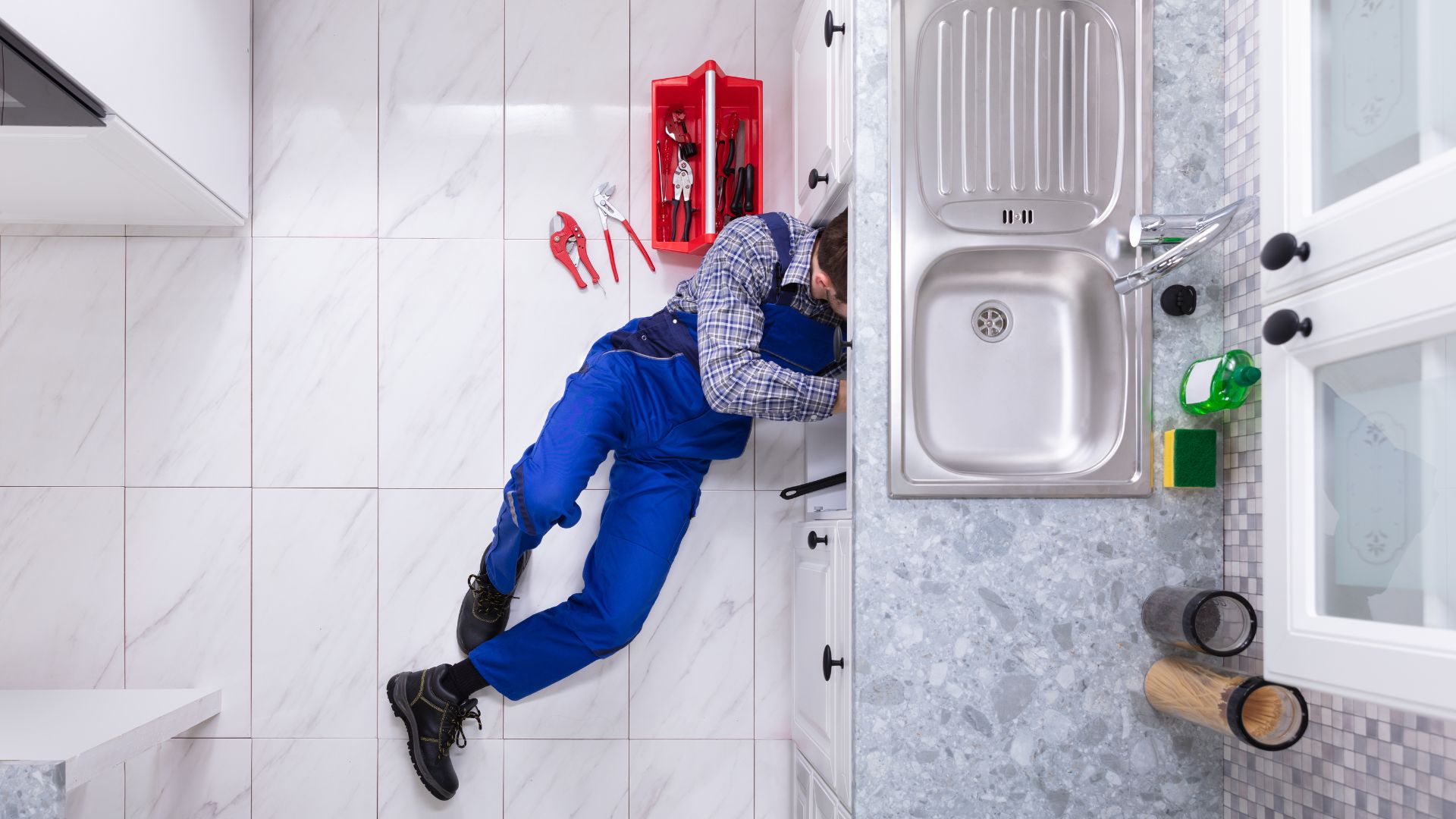 A plumber in overalls lies on the floor fixing pipes under a kitchen sink, with tools scattered nearby.
