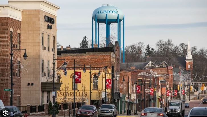 A view of a small town street with storefronts, festive banners, and a large water tower with "albion" written on it in the background.