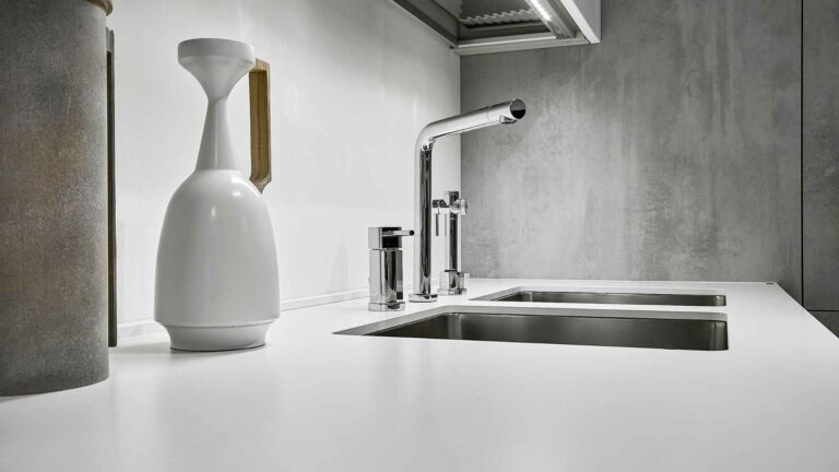 Minimalist kitchen counter featuring a modern faucet that exemplifies how to choose the right faucet for your kitchen or bathroom. Equipped with a dual sink, white bottle, metallic containers, and a grey backsplash.