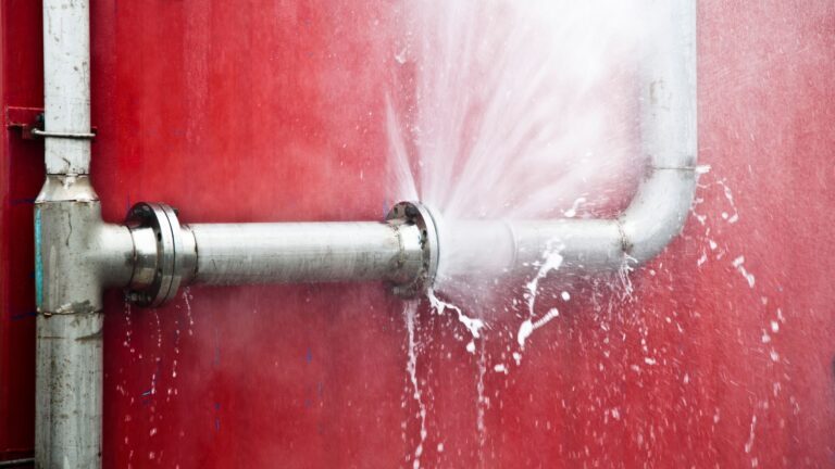 A burst metal pipe sprays water forcefully against a red wall