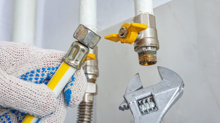 A gloved hand holds a gas hose near a metal connector and valve, with an adjustable wrench