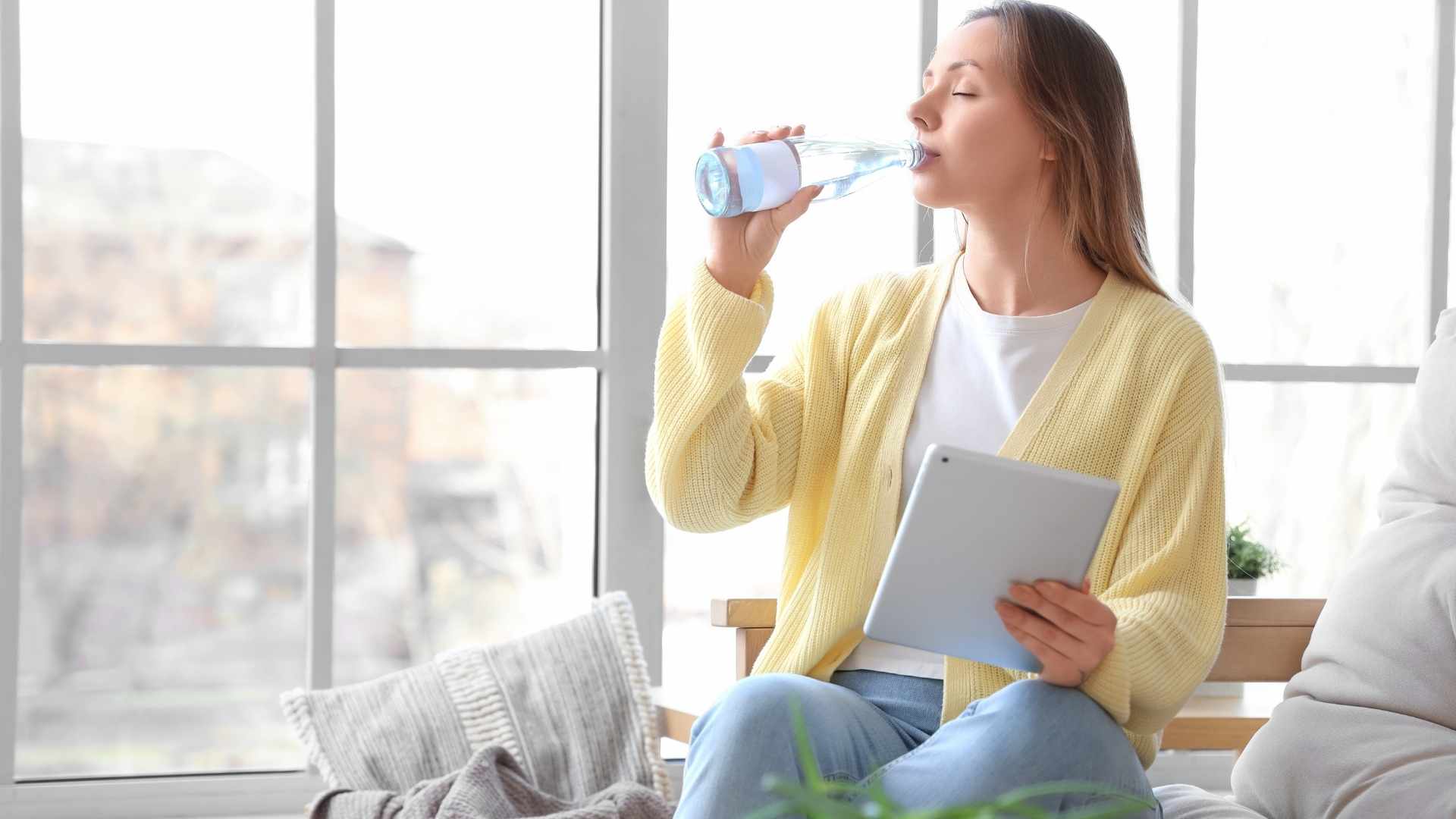 A woman sits by a window, holding a tablet & water bottle