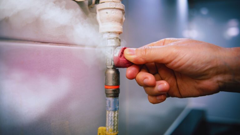 A hand turning a valve on a gas line with steam or gas leaking from the connection point, indicating a possible Gas Line Leak.