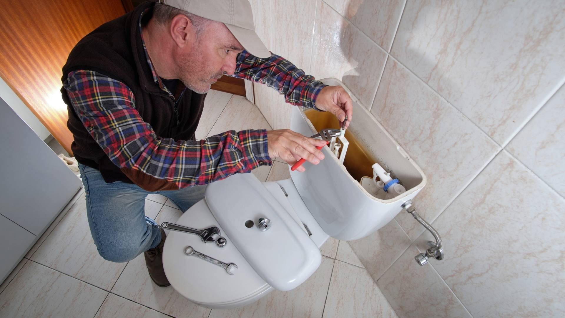 toilet plumbing in michigan A man wearing a cap and plaid shirt uses pliers to repair the inside of a toilet tank in a tiled bathroom.