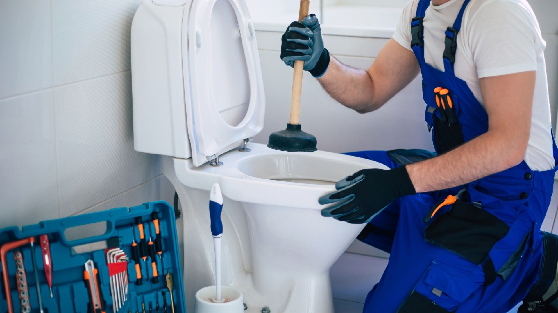 A plumber in blue overalls uses a plunger on a toilet in a bathroom, with a toolbox and cleaning brush nearby—ready to handle any plumbing issue, including gas line leaks.