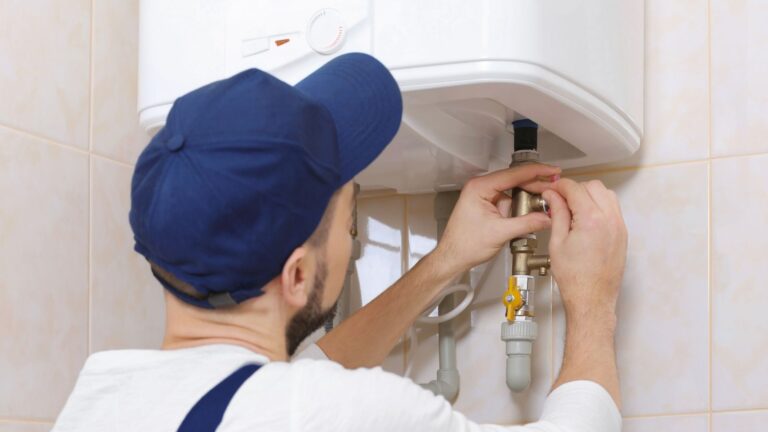 water heater replacement in michigan Person in a blue cap and white shirt adjusting pipes under a wall-mounted water heater in a tiled room.