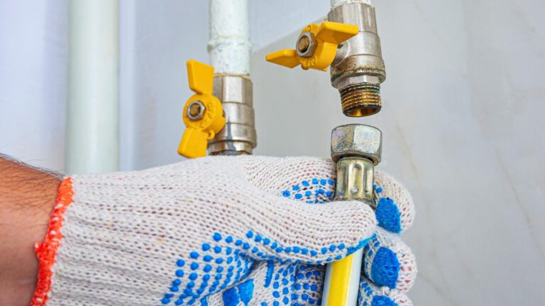 gas line repair in michigan A gloved hand holds a flexible gas hose near a shut-off valve with a yellow handle, preparing to connect or disconnect it.