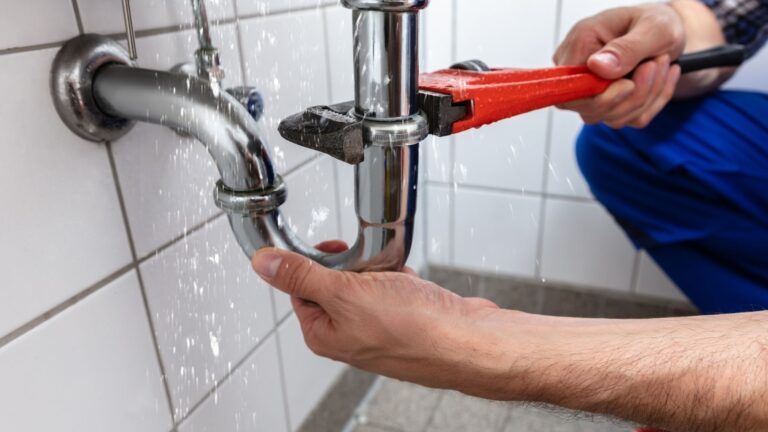 plumbing services in jackson Person using a pipe wrench to fix a leaking metal pipe under a sink, with water spraying from the joint.