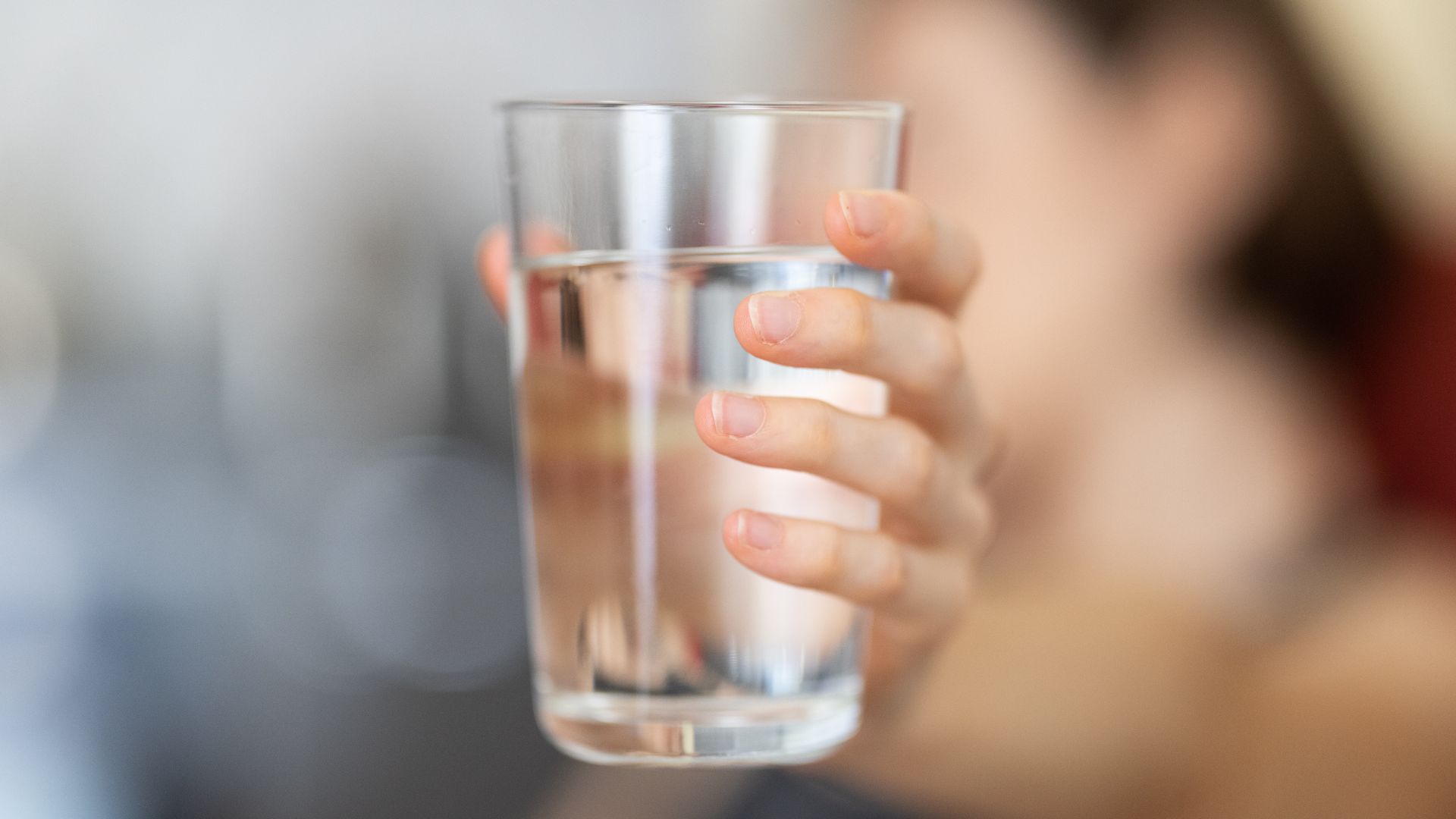 A person holds a clear glass of water, with their hand and fingers visible around the glass.