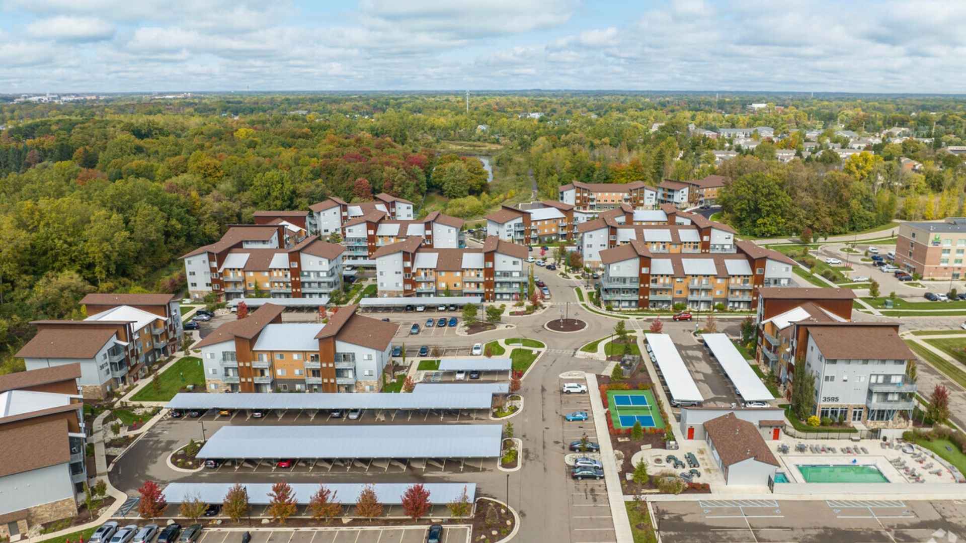Aerial view of a modern apartment complex with multiple buildings, parking areas, trees, and recreational facilities, surrounded by greenery under a partly cloudy sky.