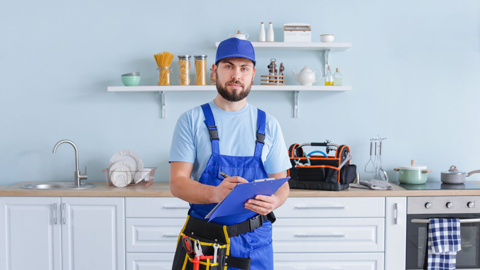 A repairman in blue overalls and cap holds a clipboard in a kitchen.